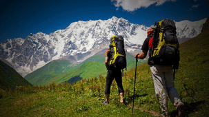2 hikers carrying large backpacks trekking along a grassy trail toward a majestic, snow-covered mountain peak.