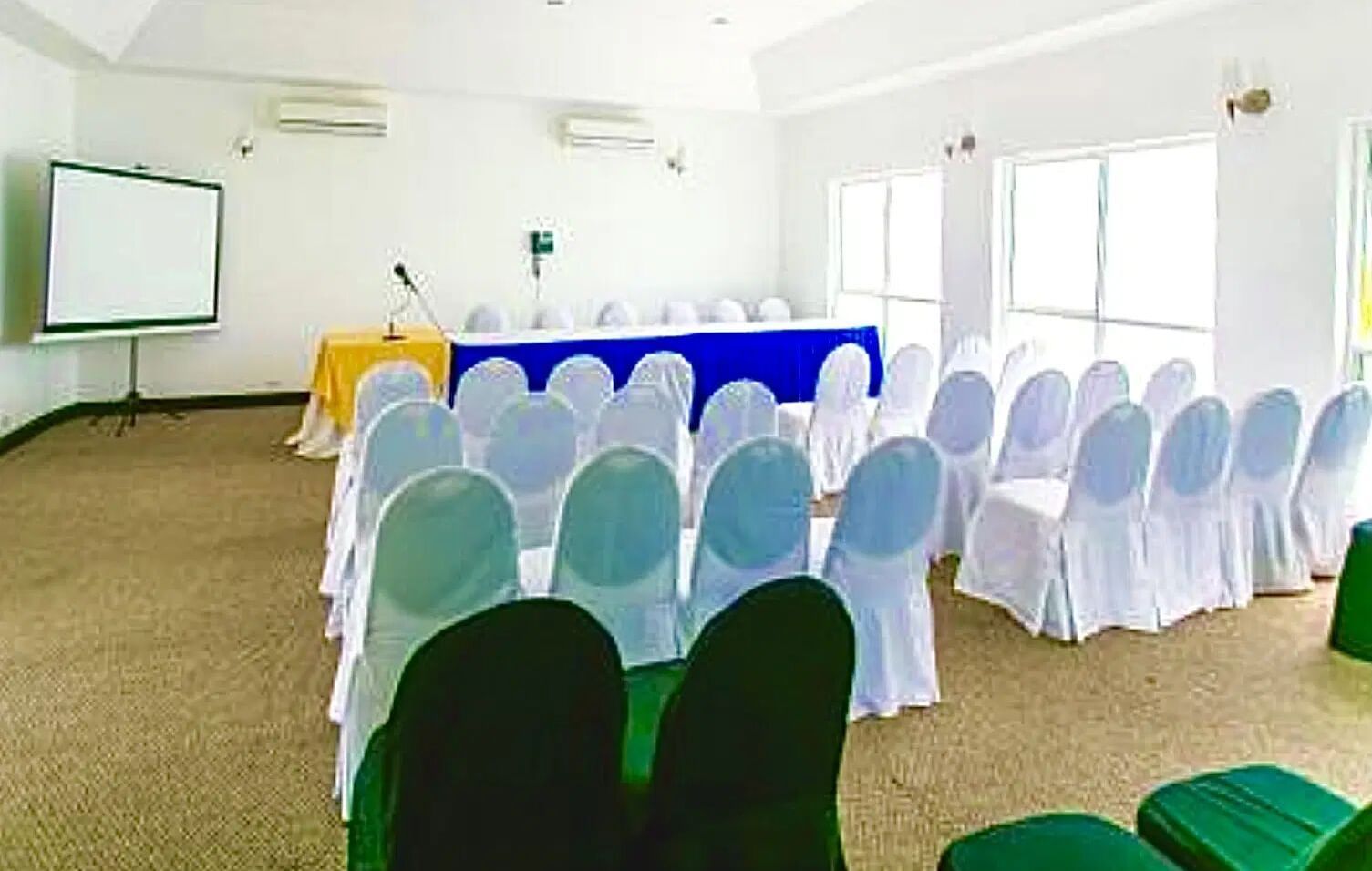 A meeting room is set up with rows of white chairs facing a table with a white tablecloth and a projector screen | Karnak | Altamont Court, Jamaica