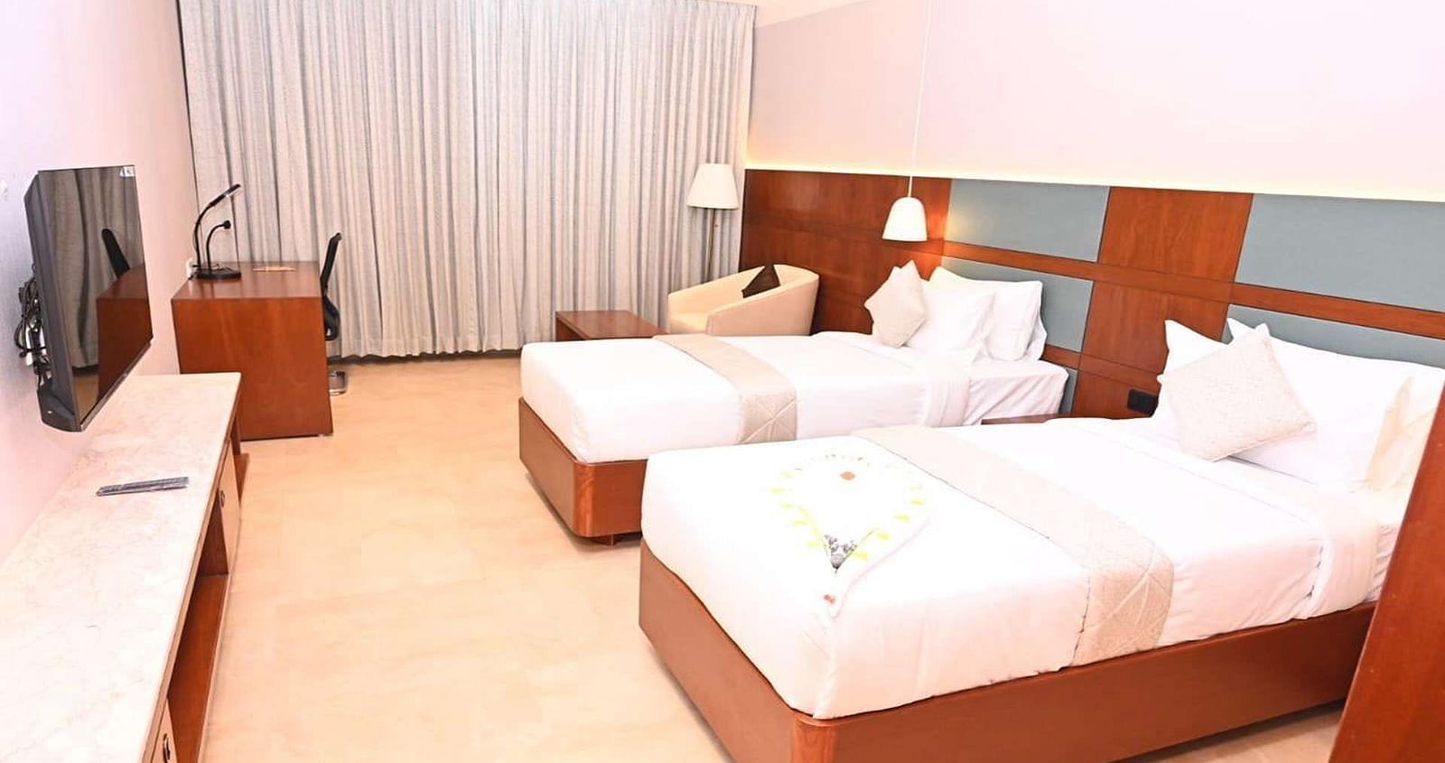 A Executive Suite at Dhanalakshmi Srinivasan Hotels, Perambalur featuring a neatly made twin beds with white linens and pillows, a wooden headboard, and a desk with a chair.