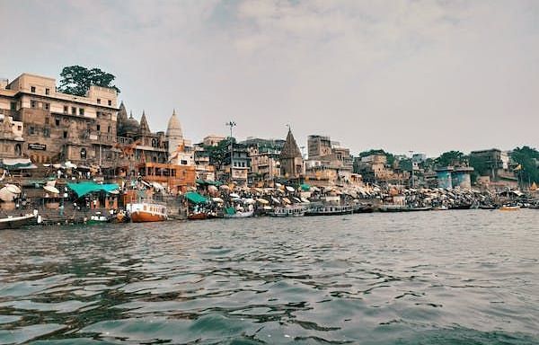 A bustling area beside a river body is alive with activity, featuring colorful boats bobbing in the water and charming houses lining the banks. The scene captures the vibrant energy of community life, with people engaging in various activities, creating a 