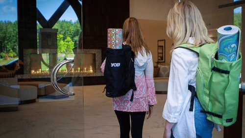 2 women with backpacks walk into a modern, spacious lobby with large windows and a glowing fireplace at YO1 Longevity and Health Resorts