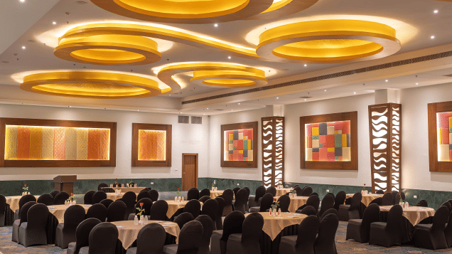 A spacious banquet hall featuring round tables with black chair covers, modern ceiling lights in golden tones and colourful wall art, set within the best wedding resort in Udaipur—Bamboo Saa Resort & Spa.