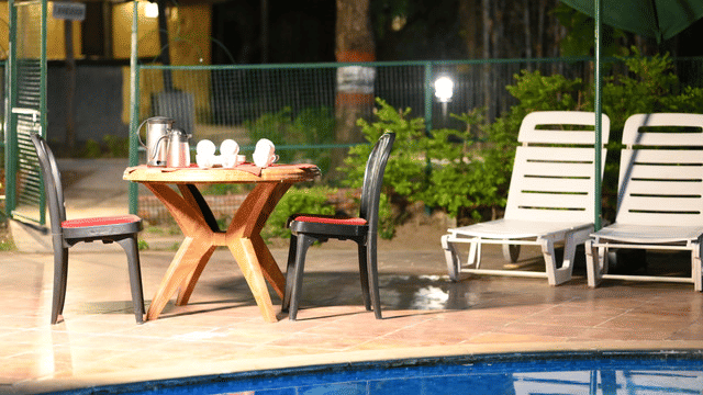 The poolside area with a table setup, chairs, lounge beds, and a glass partition next to the swimming pool at Infinity Resorts Bandhavgarh.