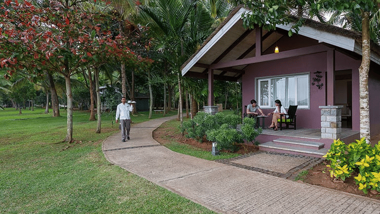 Waterfront Villa at The Serai Kabini featuring a staff member walking on the pathway surrounded by verdant manicured lawn and a couple of guests on the patio