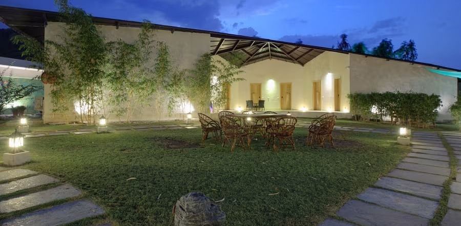 A building with lit windows at dusk or dawn and has a modern or minimalist architectural style. A paved pathway leads towards the building, and greenery around - Bamboo Saa Resort & Spa, Udaipur