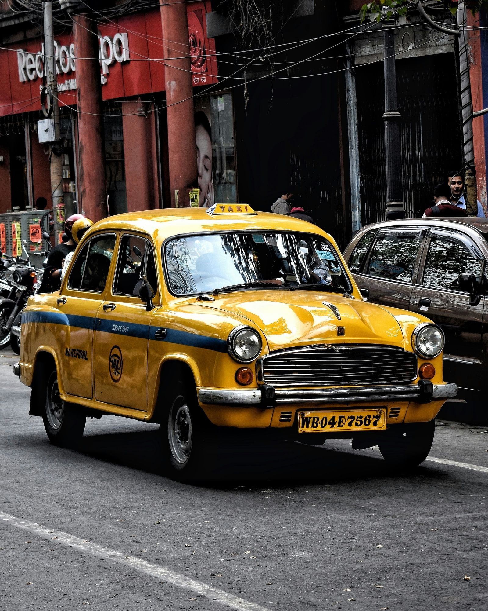 A classic taxi with a light on top drives on a city street with buildings in the background.