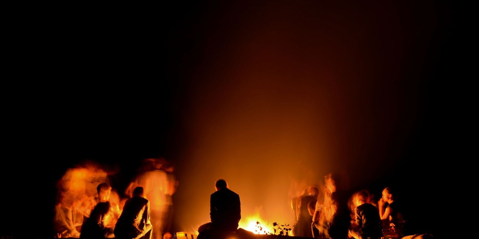 A silhouette of people gathered around a large, roaring bonfire at night, with an intense glow lighting up the scene against the darkness.