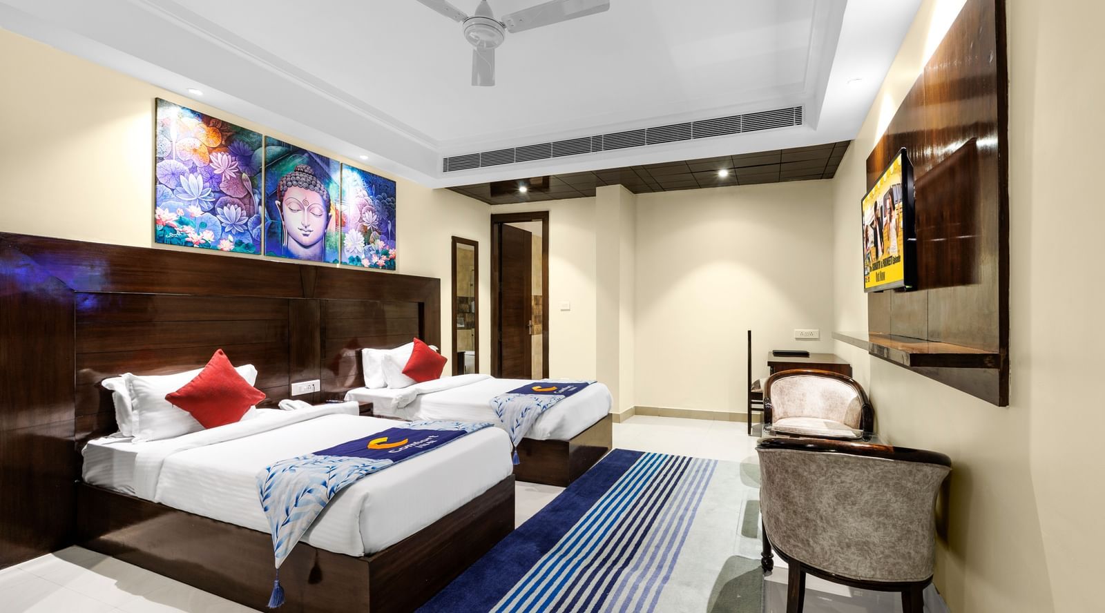 Elegant room with twin beds, ceiling fan, rug, chairs, wall-mounted TV, and colourful artwork above the beds at Inde Reva Resorts Rishhikesh.