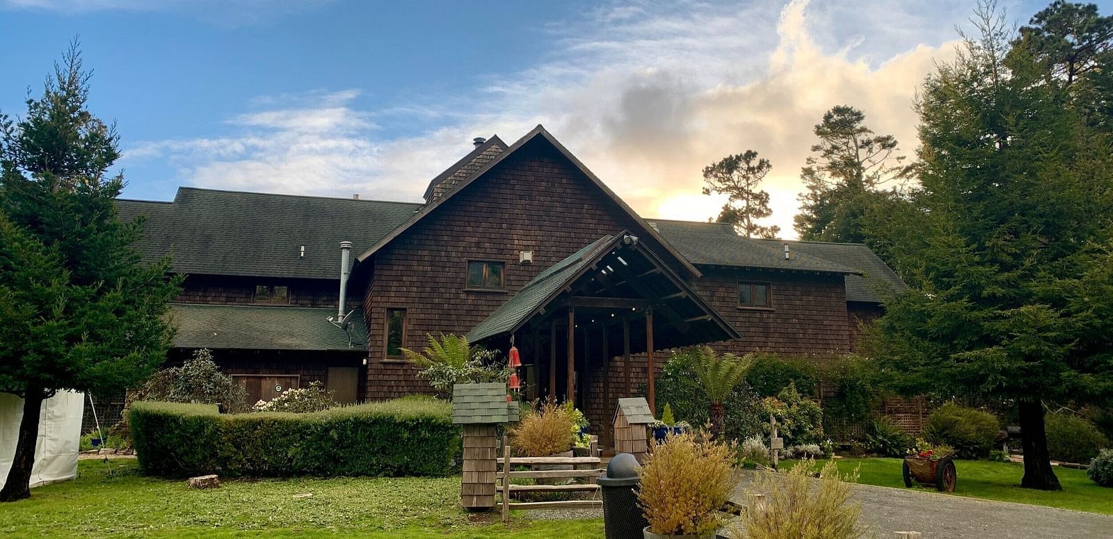 An image of cottage style house surrounded by trees and there is also a lawn in the front yard