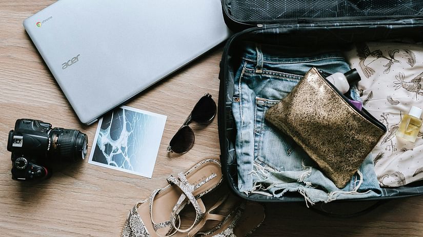 A suitcase packed with clothes, a book, and sunglasses sits next to a laptop.