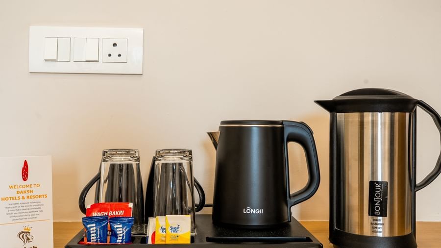 A tray kept on a table at Daksh The Grand Gowardhan, Dwarka, with a kettle, cups, and tea/coffee supplies with an electrical socket beside it.
