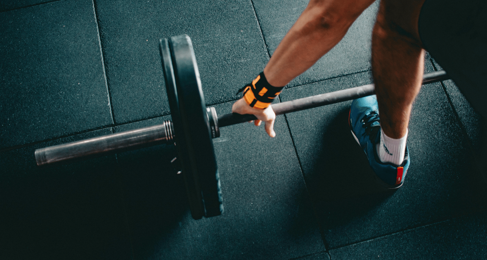 A close-up view of person's hand wearing wrist straps, gripping a metal barbell on a gym floor.