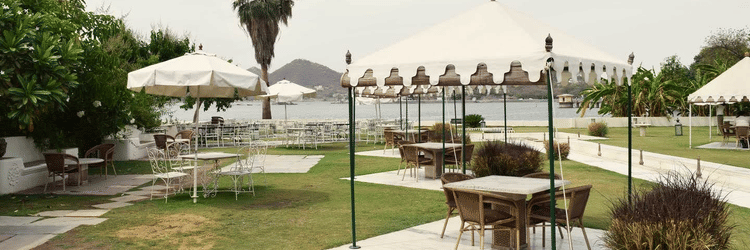 Outdoor seating area at Ram Pratap Palace, Udaipur with canopies, tables, chairs, and greenery, overlooking a lake with a palm tree and hills in the background.