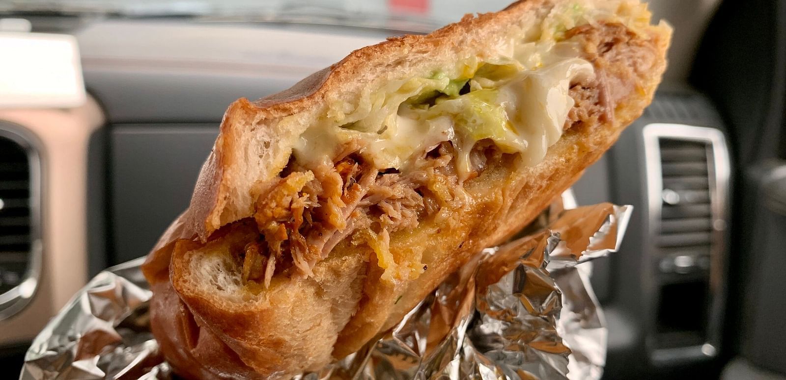 A close-up shot of a partially eaten sandwich reveals shredded meat, melted cheese, and shredded lettuce inside a crusty roll, resting on crumpled aluminum foil.