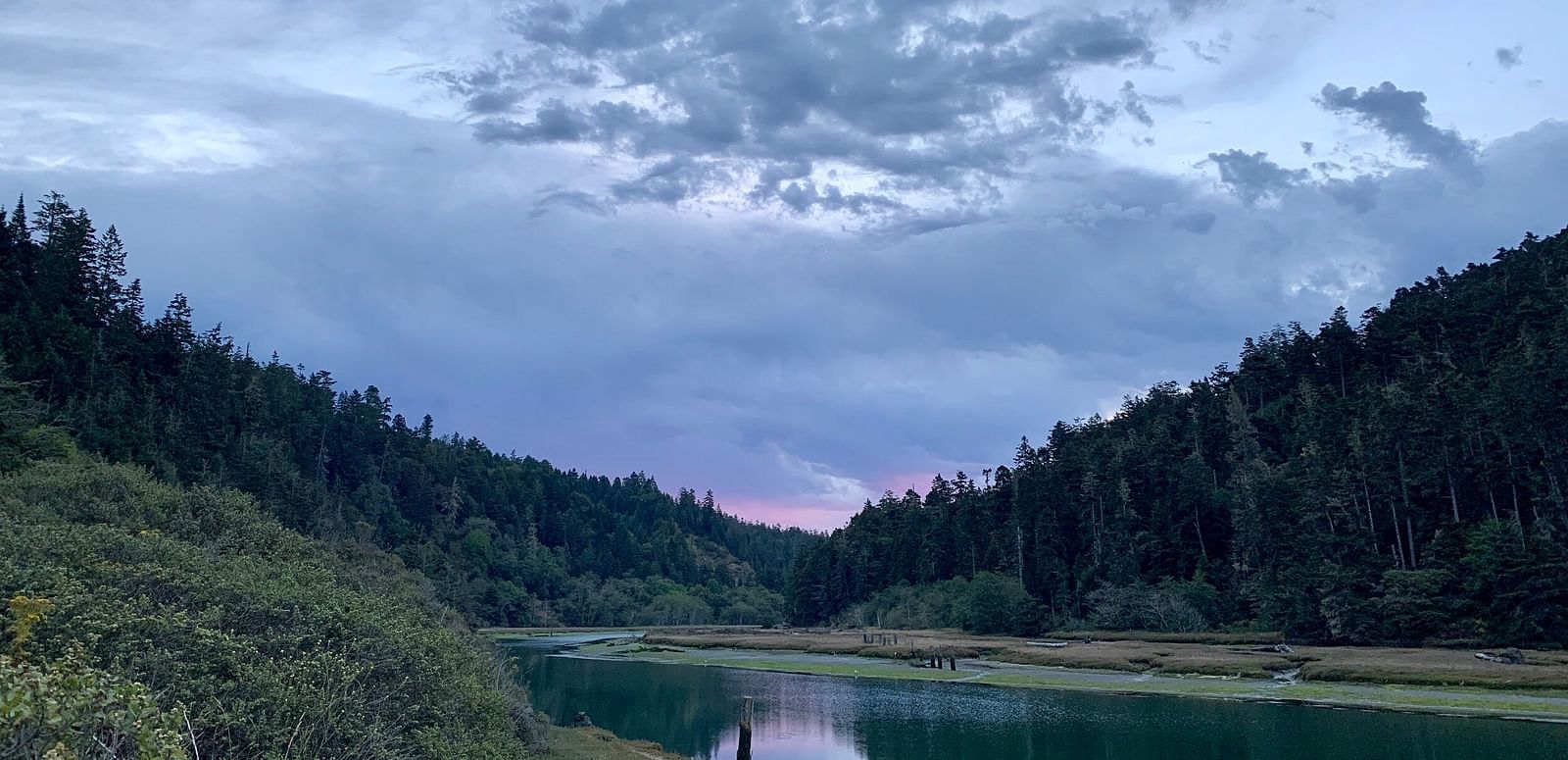 A river winds through a forested valley under a dramatic, cloudy sky at dusk.