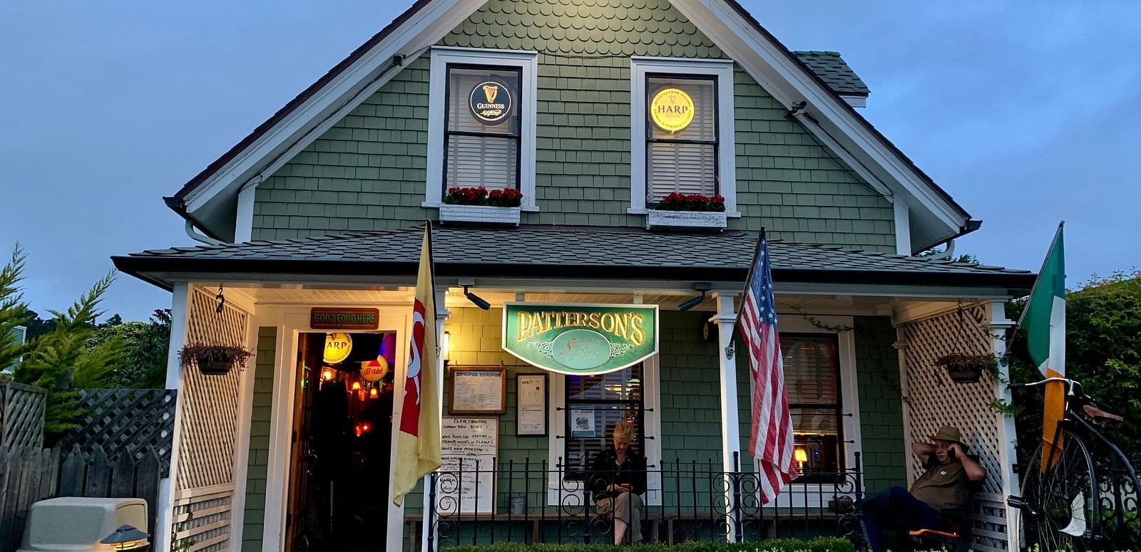 A building with a gabled roof and lit windows at dusk, featuring flags and a sign above the entrance, with a trimmed hedge in front - Brewery Gulch Inn & Spa.