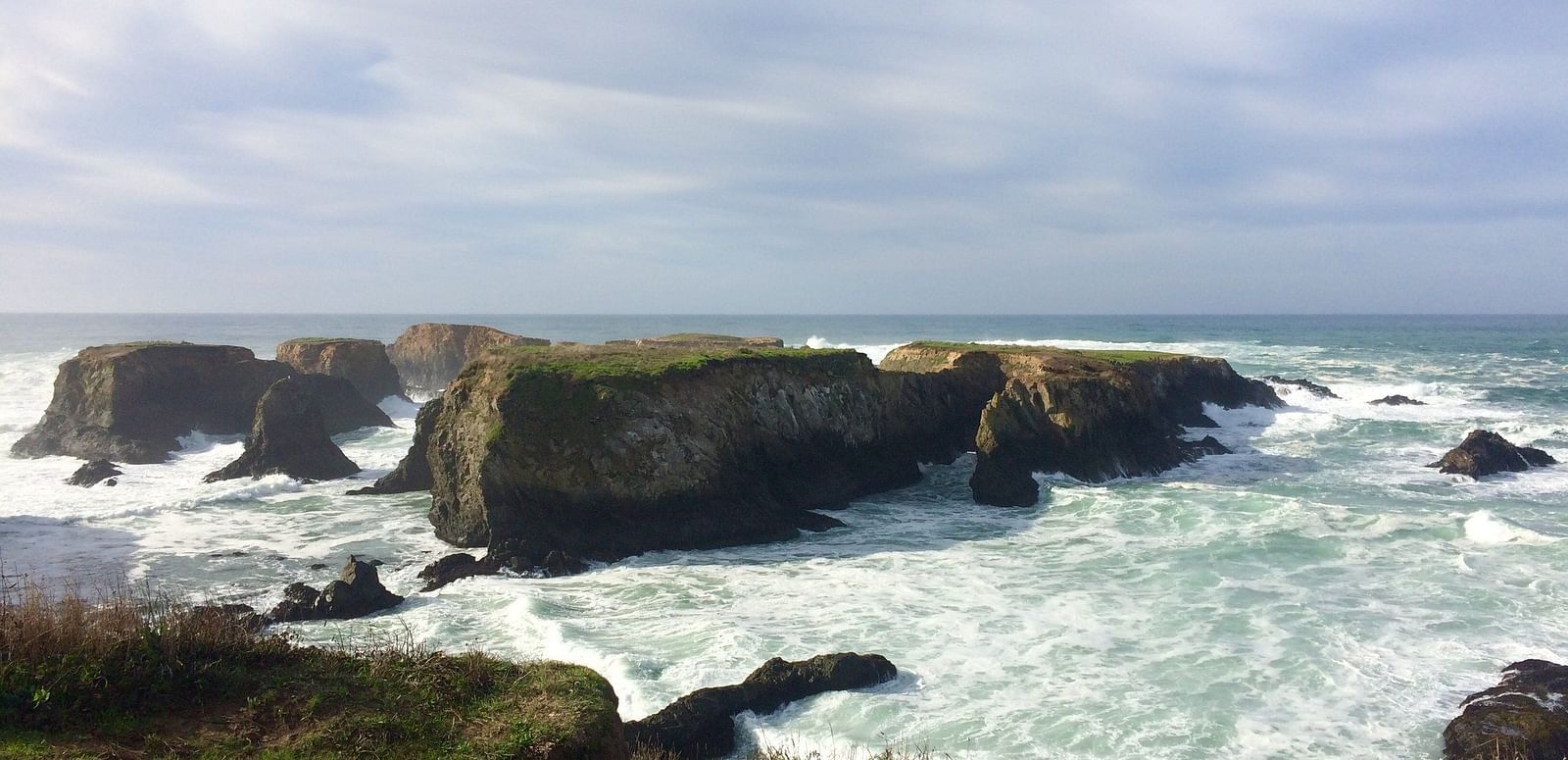 Rocky formations jut out from turbulent water, with a grassy area in the foreground under a partly cloudy sky.