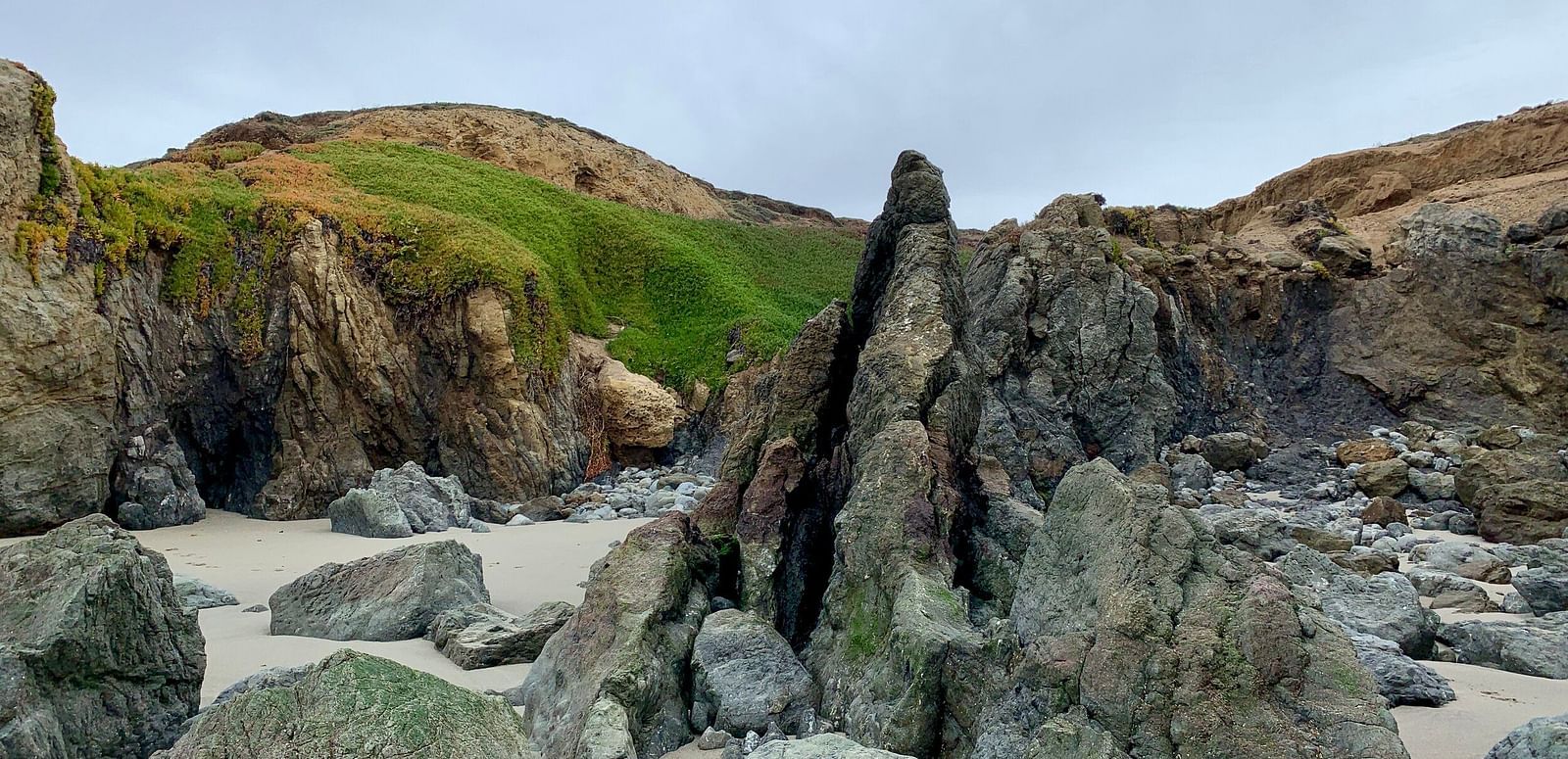 An image of a beach side featuring huge rocks and a hill beyond them