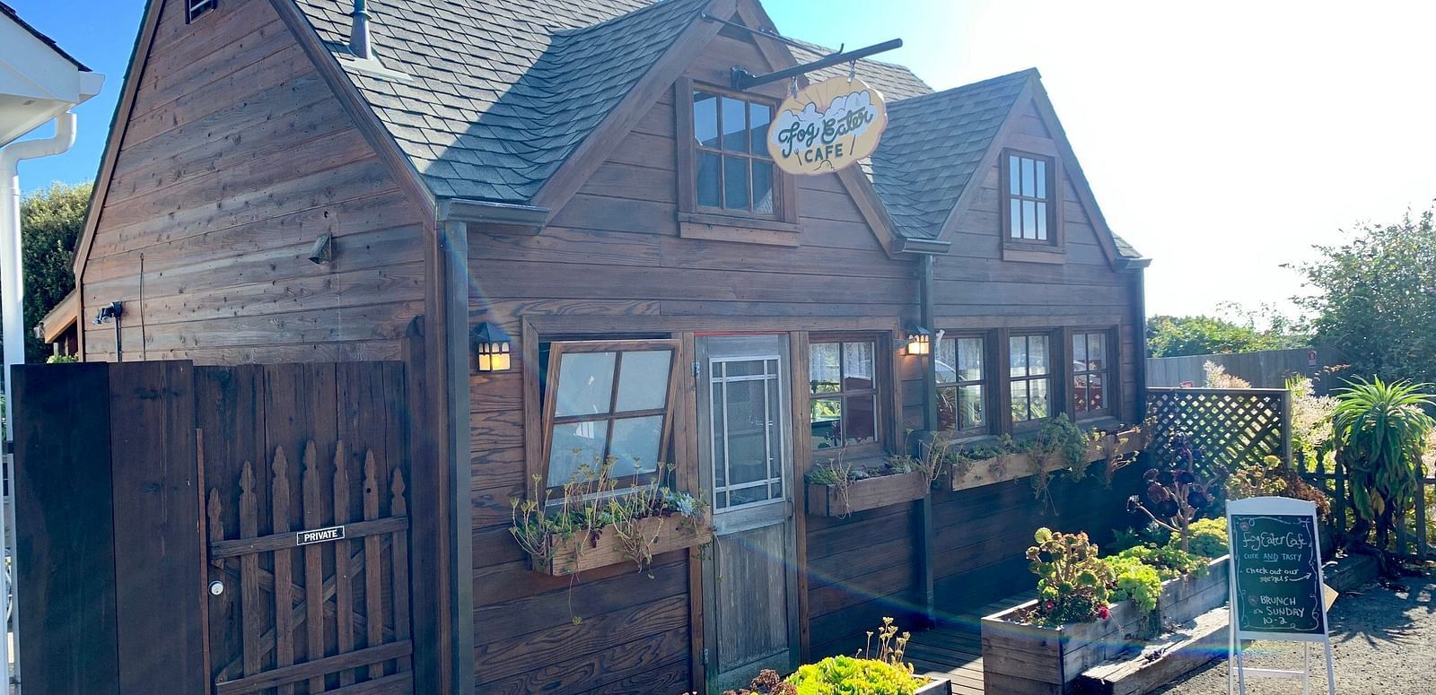 A building with a gabled roof and a sign hanging above the entrance, surrounded by a fence and potted plants.