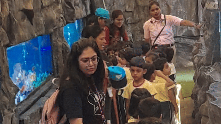 A vertical, brightly lit interior view of a crowded area at EsselWorld Fish Paradise, possibly a tunnel or hallway inside the aquarium, filled with many visitors.