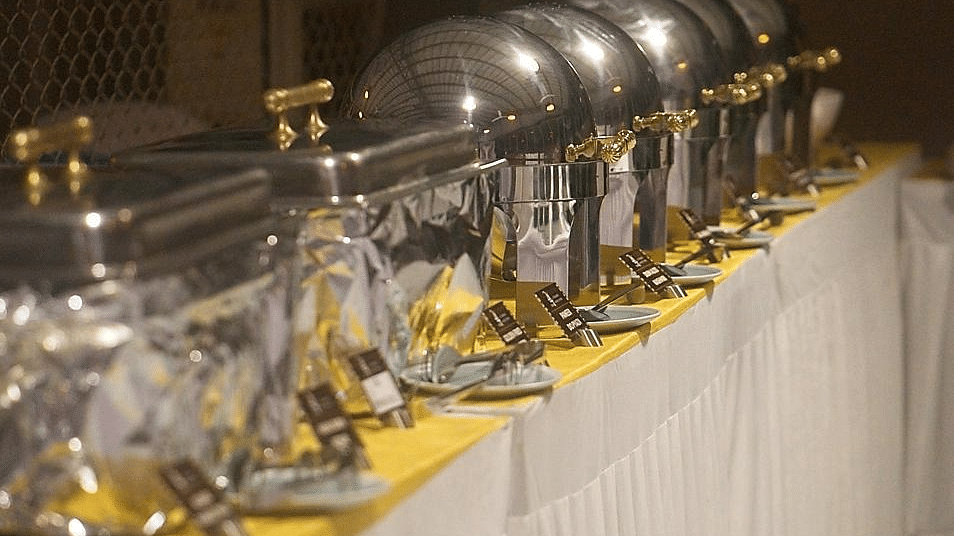 Line of chafing dishes with clear glass lids on a table with a yellow tablecloth.