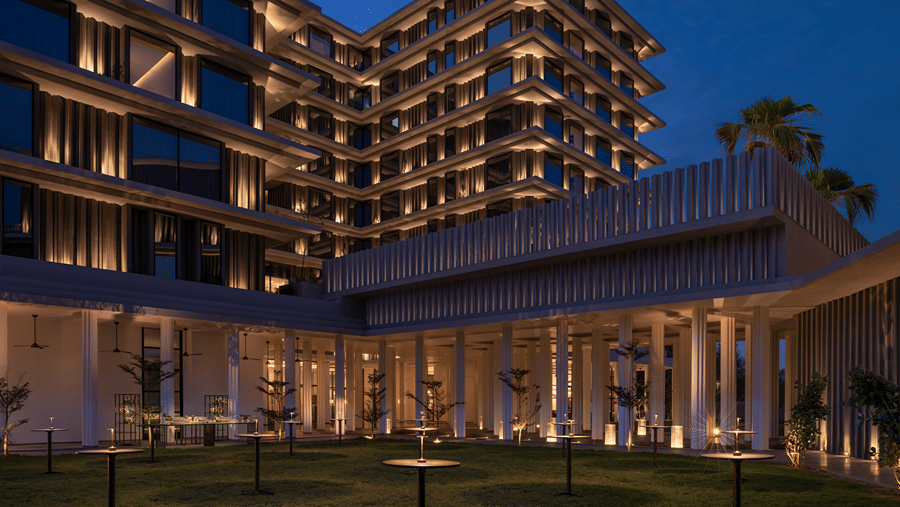 Lawn at night with lit high cocktail tables before an illuminated colonnade and a tiered hotel tower under a deep blue sky at The Residency Signature Madurai.