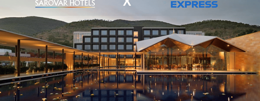 A building with several windows, restaurant with glass walls, and a corridor with plants and lights. A promotion for Amex Card holders at Sarovar Hotels.