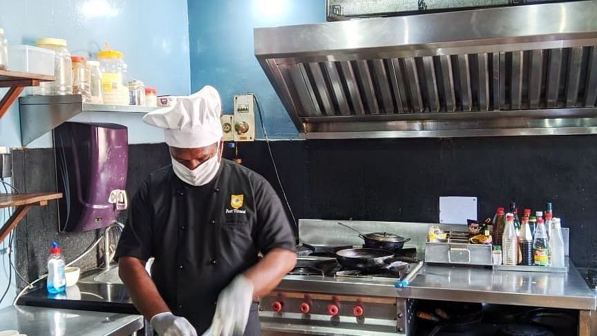 A chef wearing a black hat and coat is meticulously preparing food on a cutting board in a professional kitchen, with stainless steel surfaces and various cooking equipment visible.