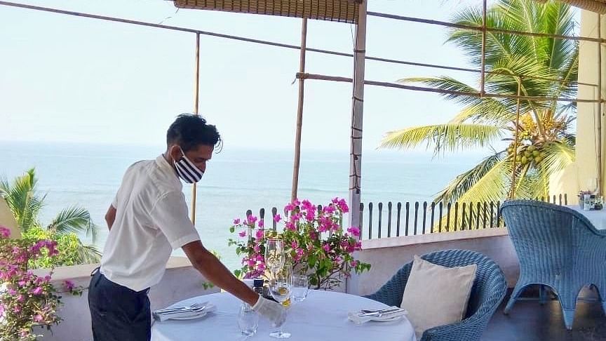 A staff member in a white uniform is setting or preparing an outdoor table at Fort Tiracol, Goa, with the ocean visible in the background through large windows or an open patio. 