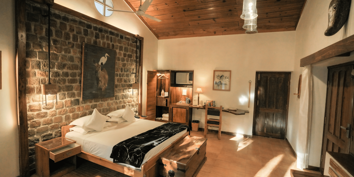 View of a room at The Den Corbett Resort and Spa Kumeria with a king bed, rustic stone wall, wooden ceiling, pendant lights, and warm ambient lighting.