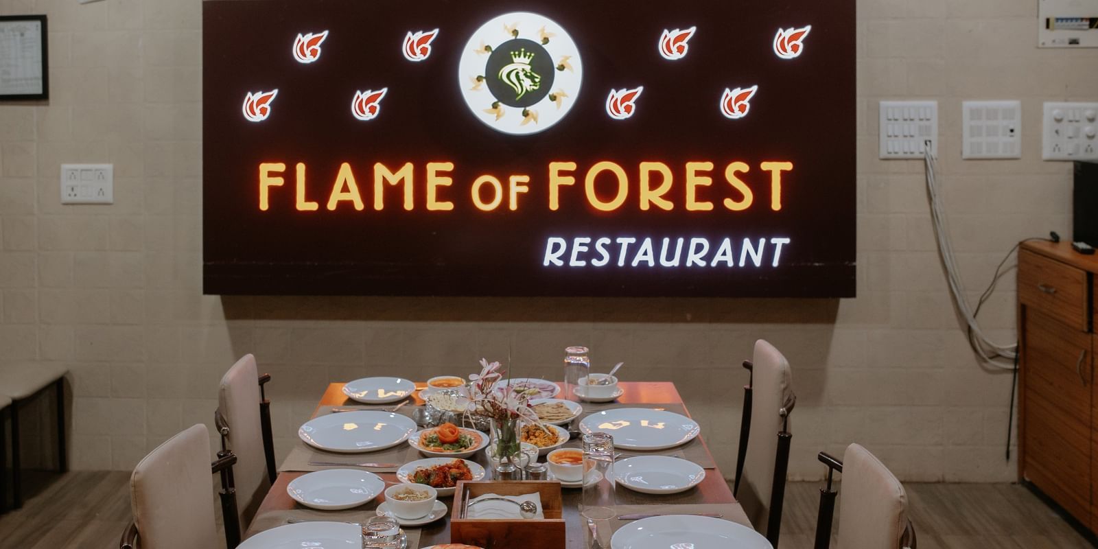 A clean, set dining table under the Flame of Forest Restaurant sign, awaiting diners, at Daksh Resort & Amusement Park, Sasan Gir.