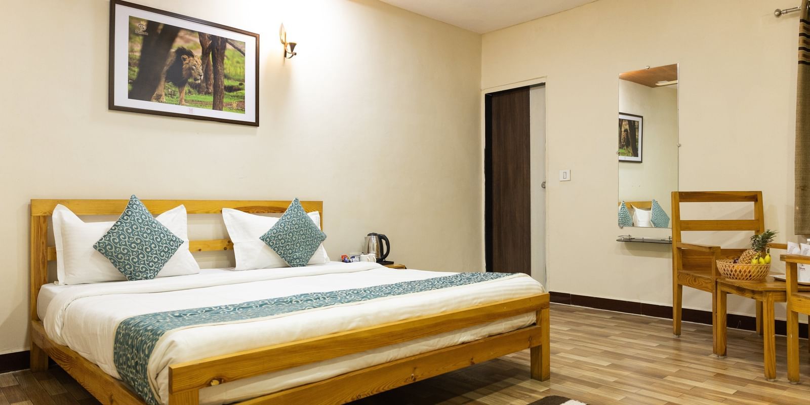 A clean room with a large bed, blue accents, a mirror, a wall art and a small chair in the corner, at Daksh Resort & Amusement Park, Sasan Gir.