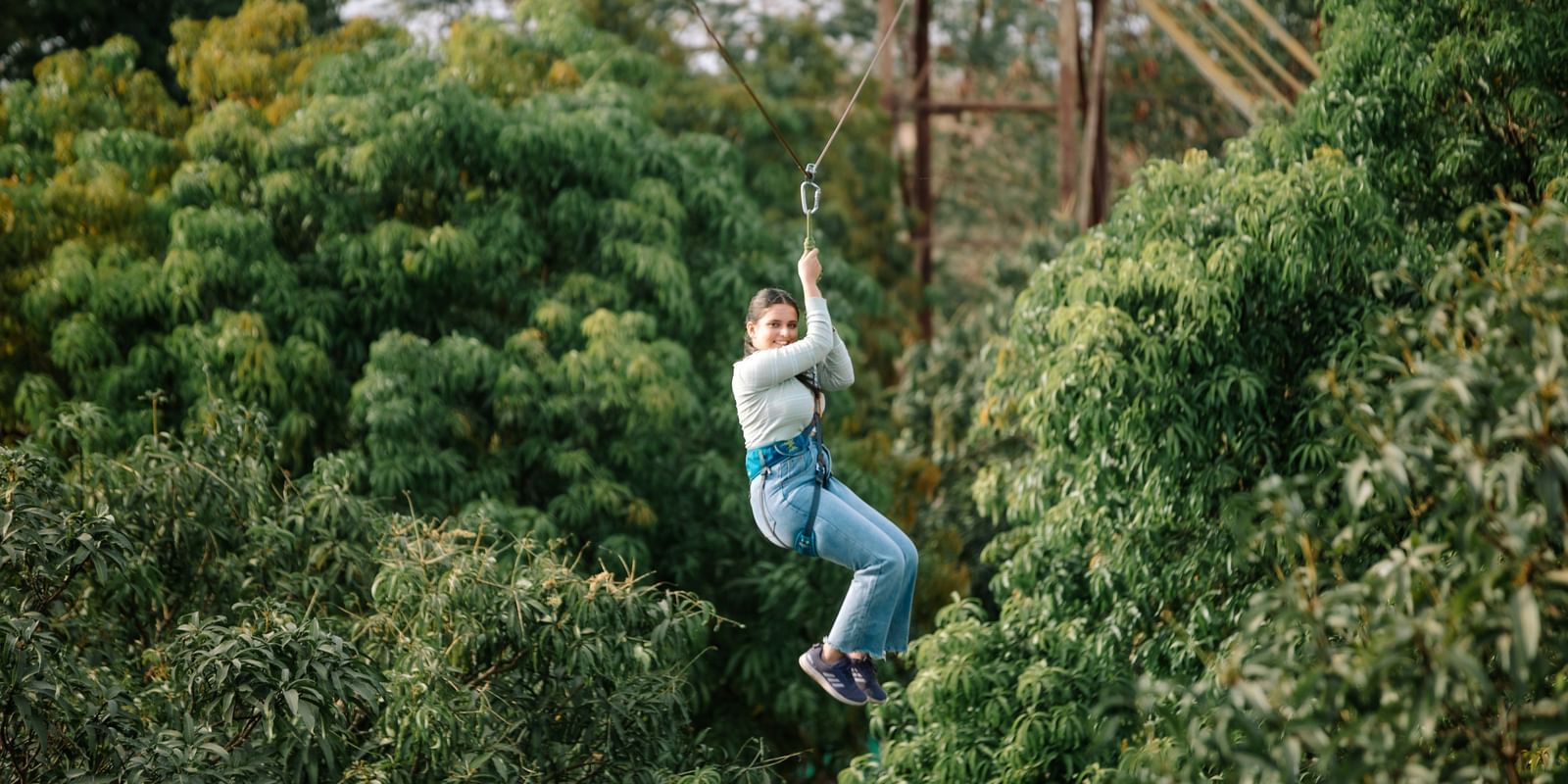 A person zip-lining through a dense green forest area, suspended mid-air by harness and cable at Daksh Resort & Amusement Park.
