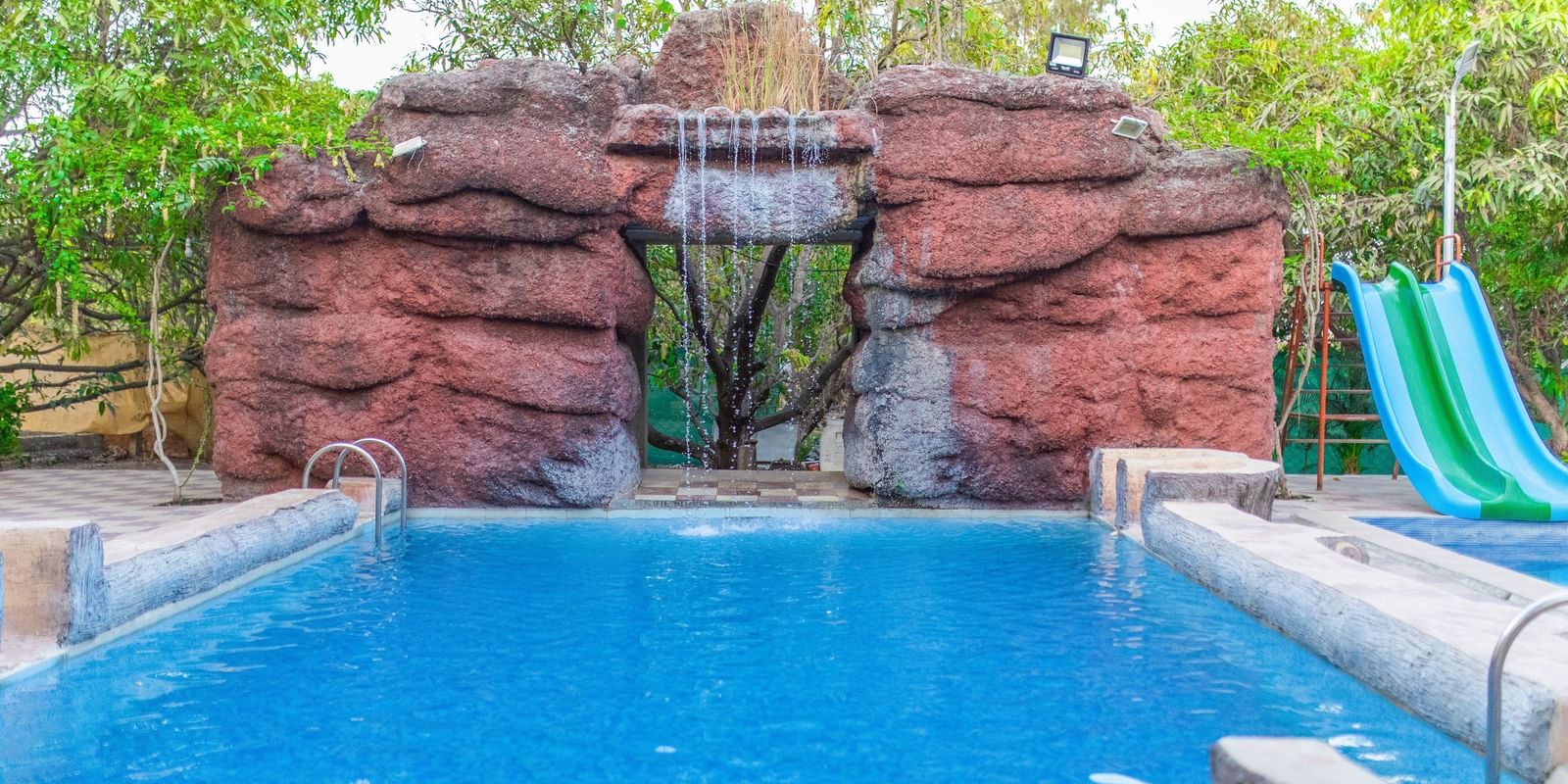 A swimming pool with a slide featuring a man-made waterfall at the background at Daksh Resort & Amusement Park.