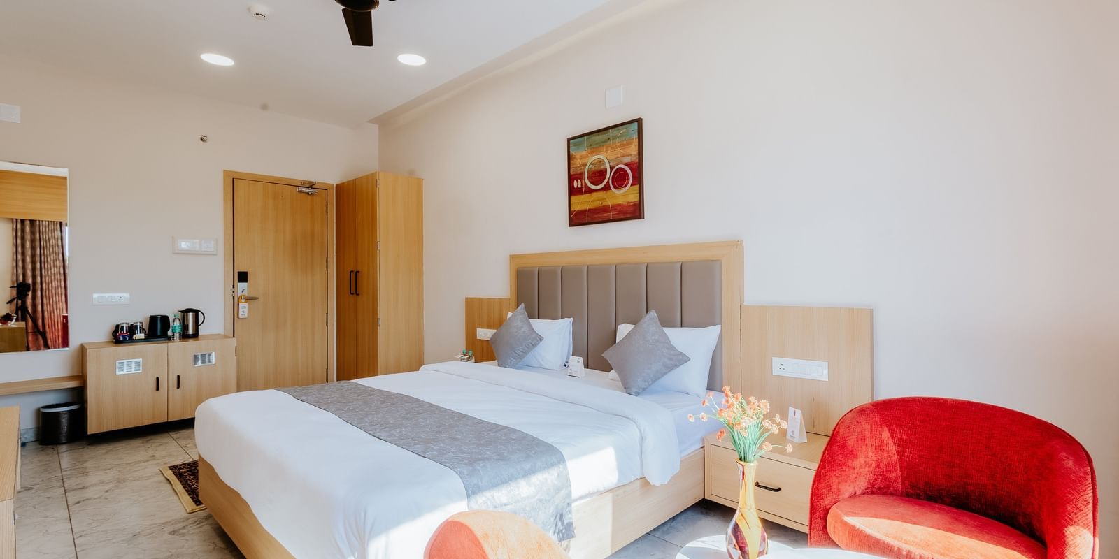Corner view of a brightly lit bedroom at Daksh The Grand Gowardhan, Dwarka, showcasing a plush double bed, a vibrant red armchair along with a coffee table in front of it.