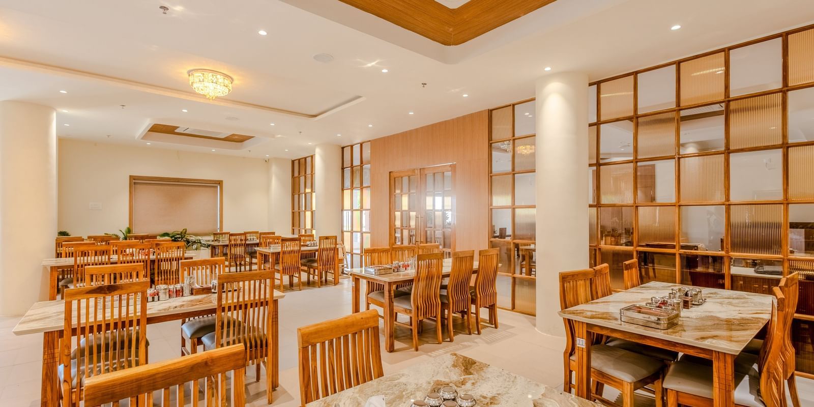 A wide view of the restaurant at Daksh The Grand Gowardhan, Dwarka, showing multiple dining tables and decorative shelving along with central air-conditioning.