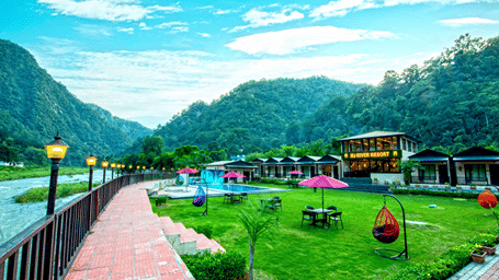 Scenic view of DLS MJ River Resort, Rishikesh, featuring a spacious lawn with outdoor seating, hanging swing chairs, a swimming pool, and resort cottages set against forested hills alongside a flowing river.