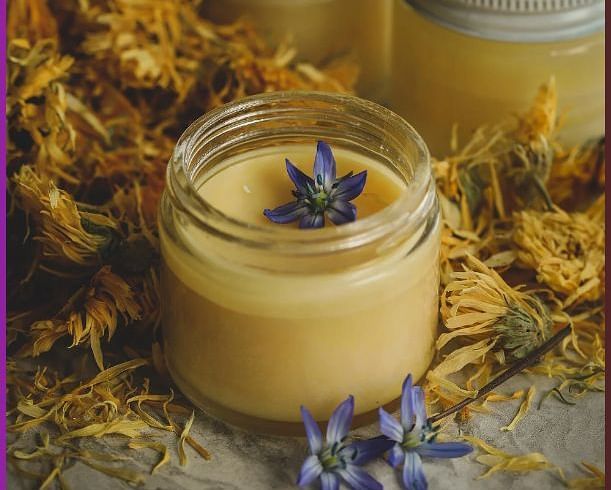 Several jars of a light-coloured, creamy substance, possibly massage cream or body butter, surrounded by dried herbs and flowers.