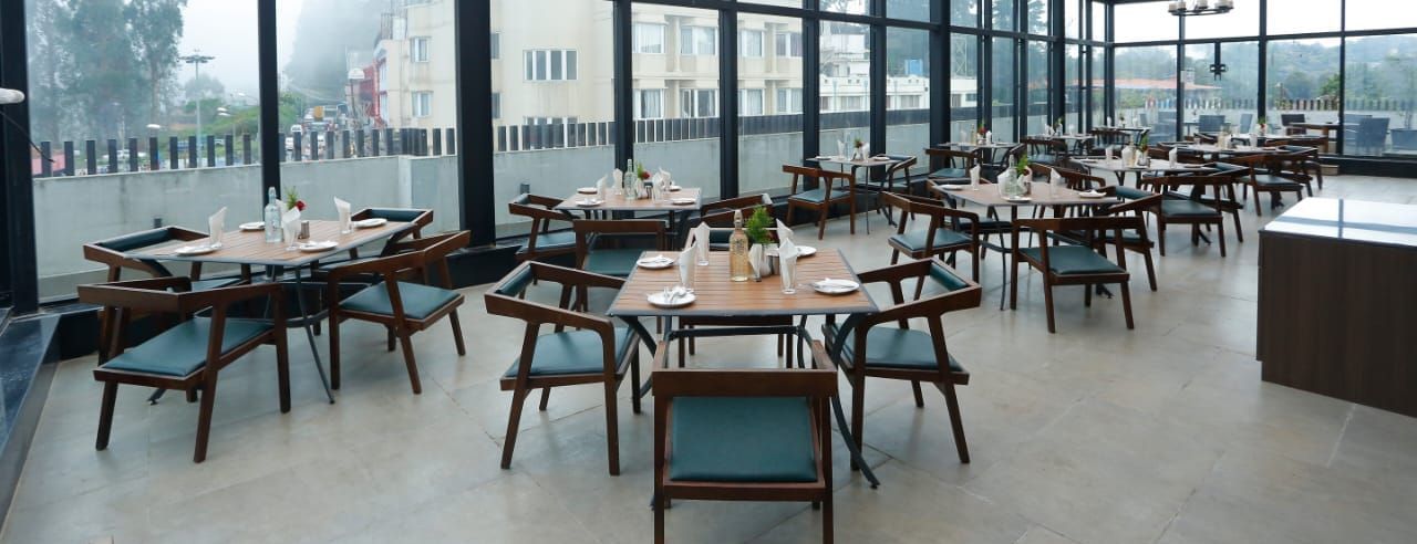 A rooftop dining area with glass walls and ceiling, offering stunning views and arranged tables and chairs for guests at The Hamuse Hotel, Kodaikanal.