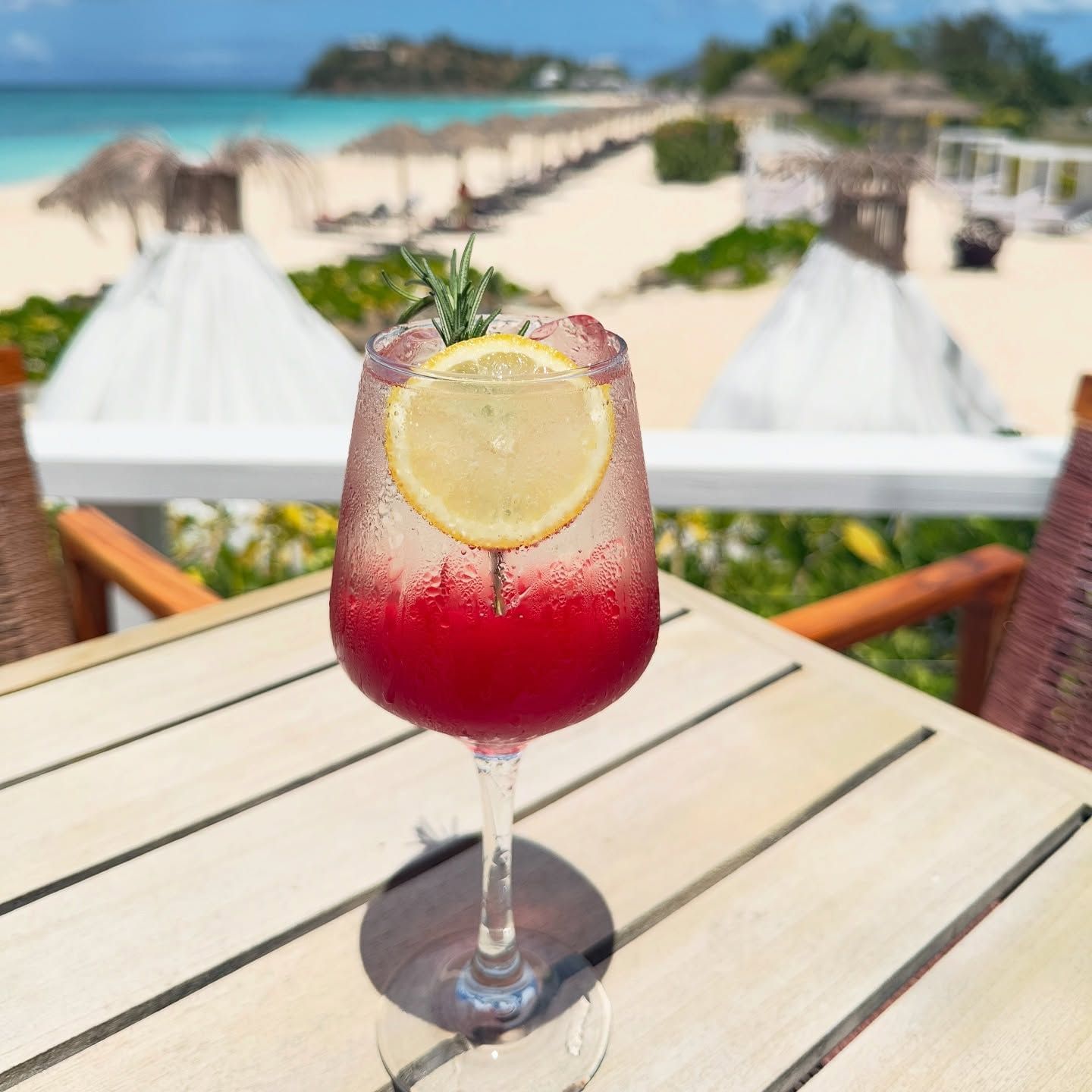 A close-up of a refreshing red drink in a glass on a wooden table, with a blurred background of a sunny beach and ocean.