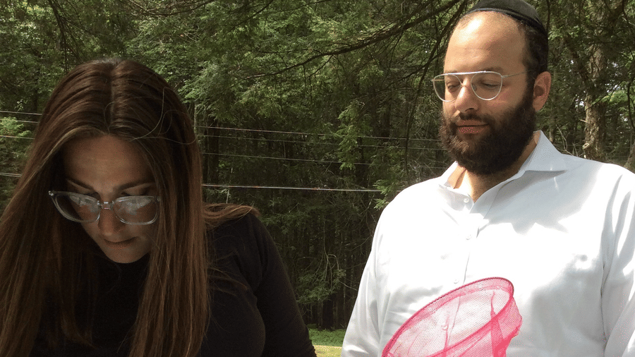 A woman examining papers on a table while holding a pink net, with a man in a white shirt standing beside her outdoors at YO1 Longevity & Health Resorts, Catskills