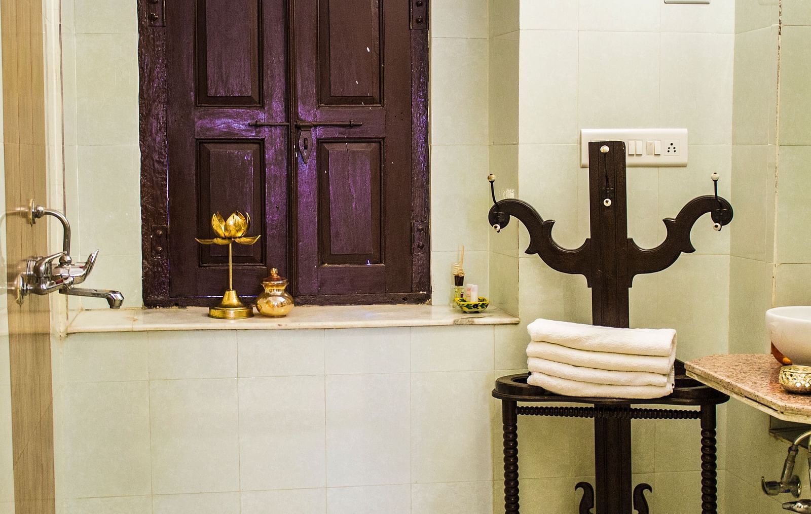 A bathroom with a dark wooden window, a towel rack with folded white towels, and a small golden lamp on the windowsill at Umaid Vilas Jaipur.