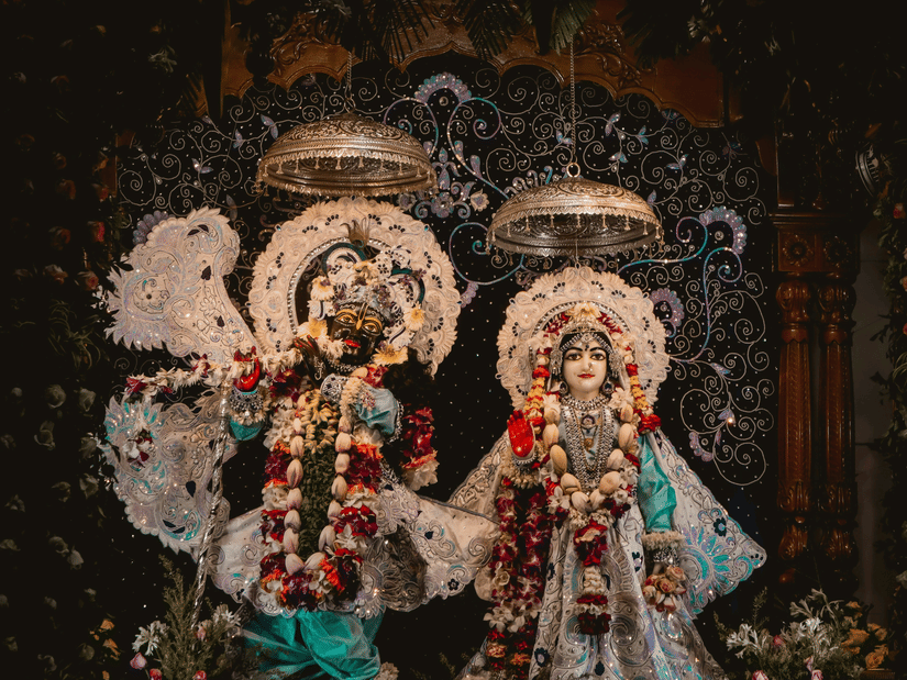 Beautifully adorned, traditional wooden deities of Radha and Krishna dressed in elegant, ornate clothing inside a dimly lit temple sanctuary.
