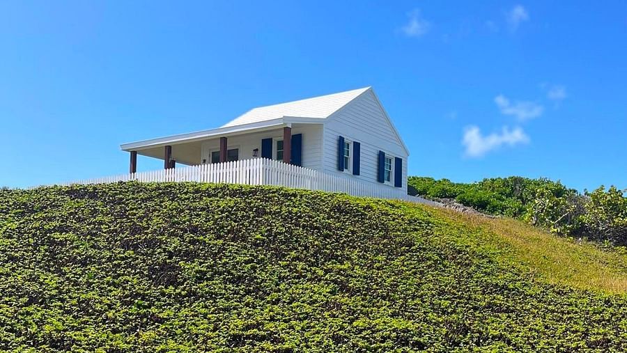  A house with a distinct roofline is situated on top of a densely vegetated, rising landscape under an open sky | Abaco Inn