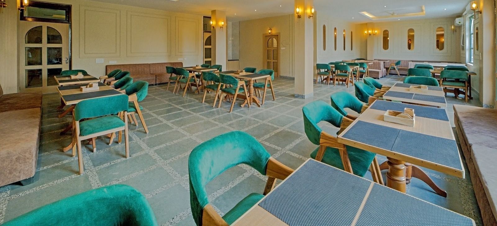 Elegant dining area at Grassfield Riveria Resort, Jaipur, with teal upholstered chairs, wooden tables, and warm lighting.