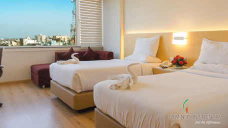 Hotel room with twin beds dressed in white linen, maroon footstools, a large window with a city view and a fruit basket on the bedside table.