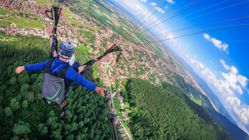 Paraglider gliding above a green valley, viewed from behind with harness lines visible, while fields and hills curve towards horizon