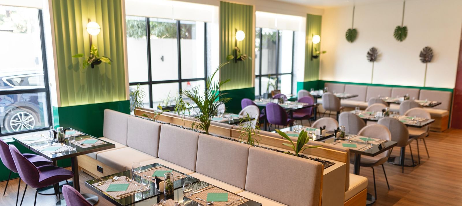 Bright, modern Meli-Melo restaurant interior with accents, large windows, potted plants, and neatly set tables with chairs and beige booth seating. Natural light fills the stylish dining area