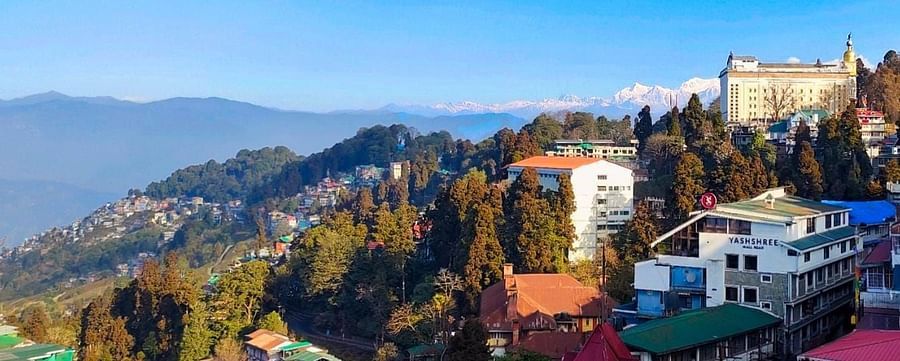 Stunning view of a hillside town with lush greenery, colourful rooftops, and snow-capped mountains in the background under a clear blue sky.