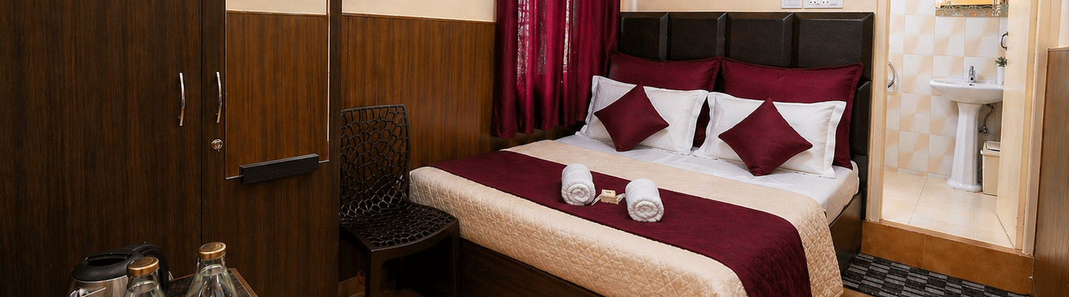 A plush bed next to a large wardrobe with a mirror, a wall-mounted fan, and a kettle setup on a table in the Deluxe Double Room at BN Tourist Home, Ooty.